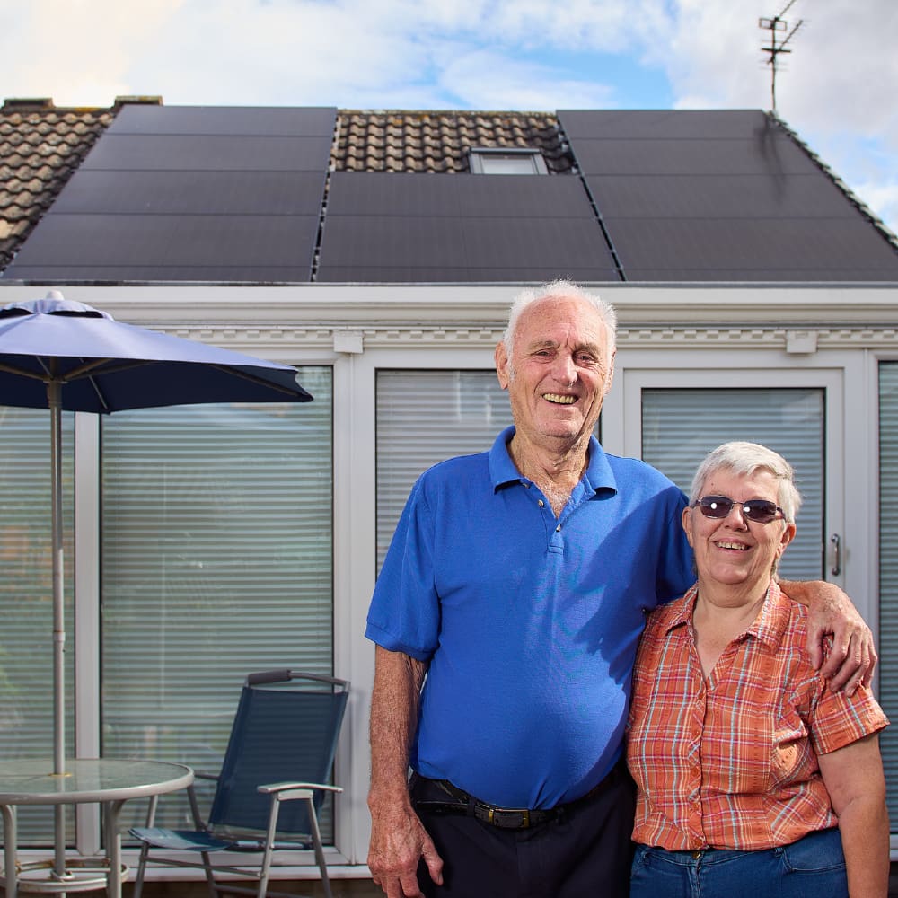 A man wearing a blue shirt stood next to a woman wearing an orange shirt. Behind them is a bungalow with solar panels on the roof. 